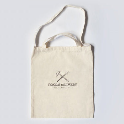 A cream-colored canvas tote bag with two top handles, featuring a black printed logo of crossed tools and the text 'TOOLS FOR LIFEBY' on the front, set against a light gray background.