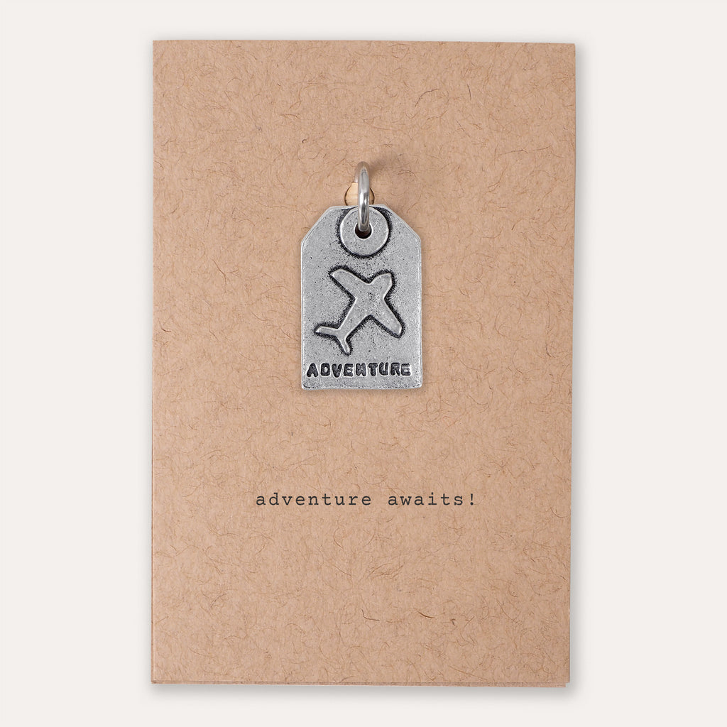 A small silver tag-shaped pendant with an airplane symbol and the word 'ADVENTURE' is attached to a kraft paper backing card that reads 'adventure awaits!'.