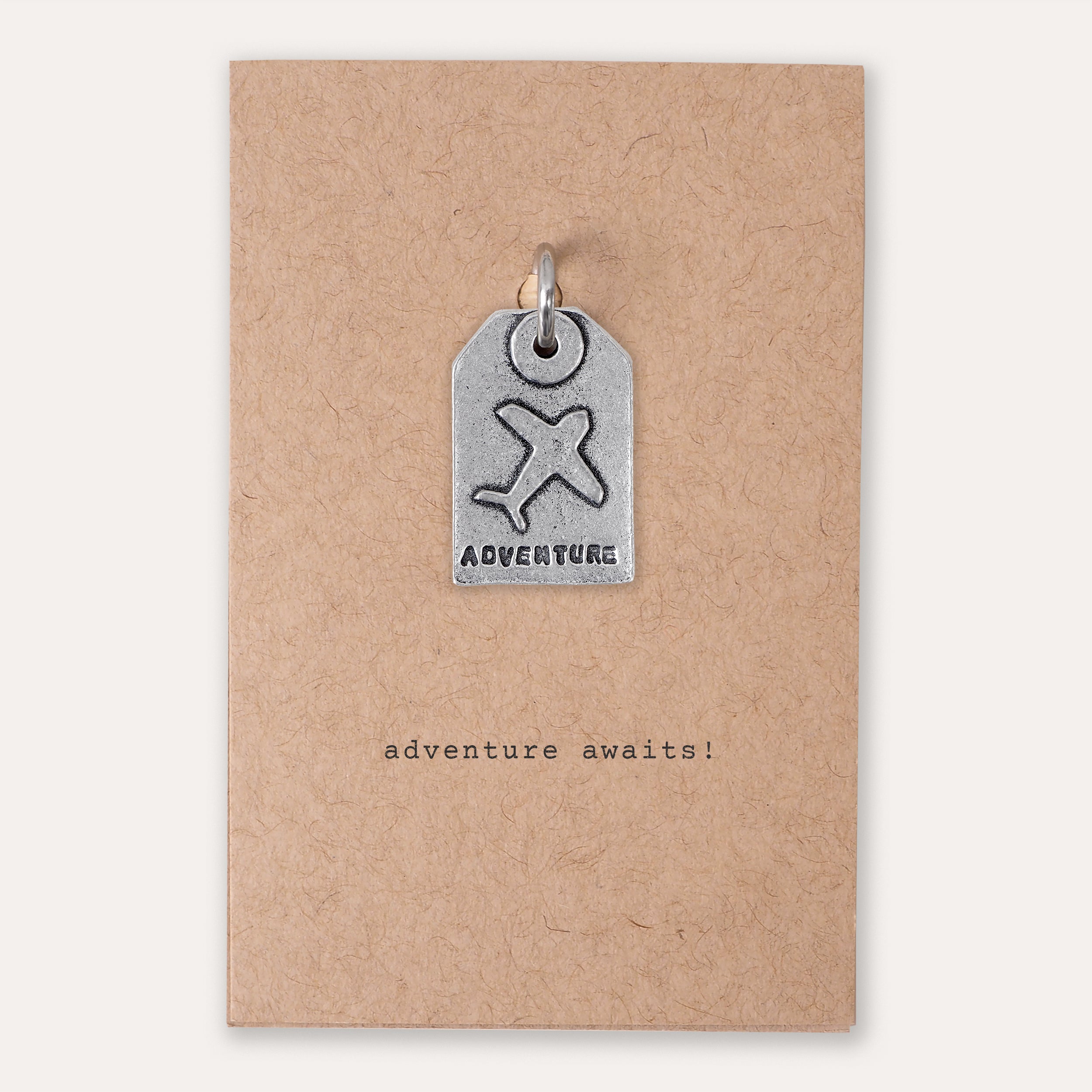 A small silver tag-shaped pendant with an airplane symbol and the word 'ADVENTURE' is attached to a kraft paper backing card that reads 'adventure awaits!'.