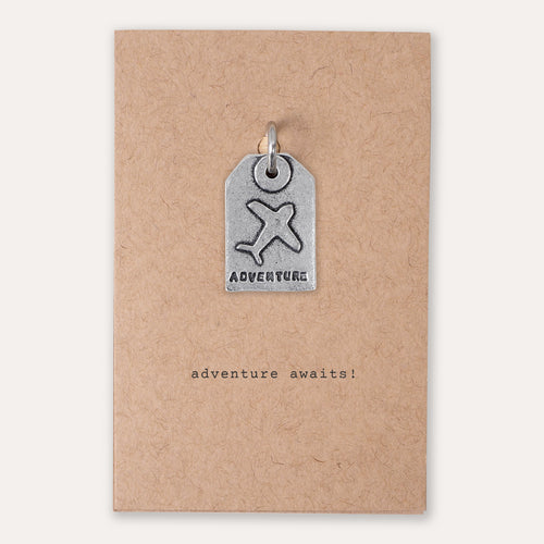 A small silver tag-shaped pendant with an airplane symbol and the word 'ADVENTURE' is attached to a kraft paper backing card that reads 'adventure awaits!'.