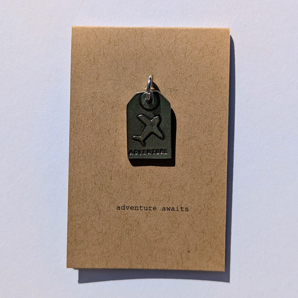 Metal plane tag shaped pendant on a brown card with 'adventure awaits' text