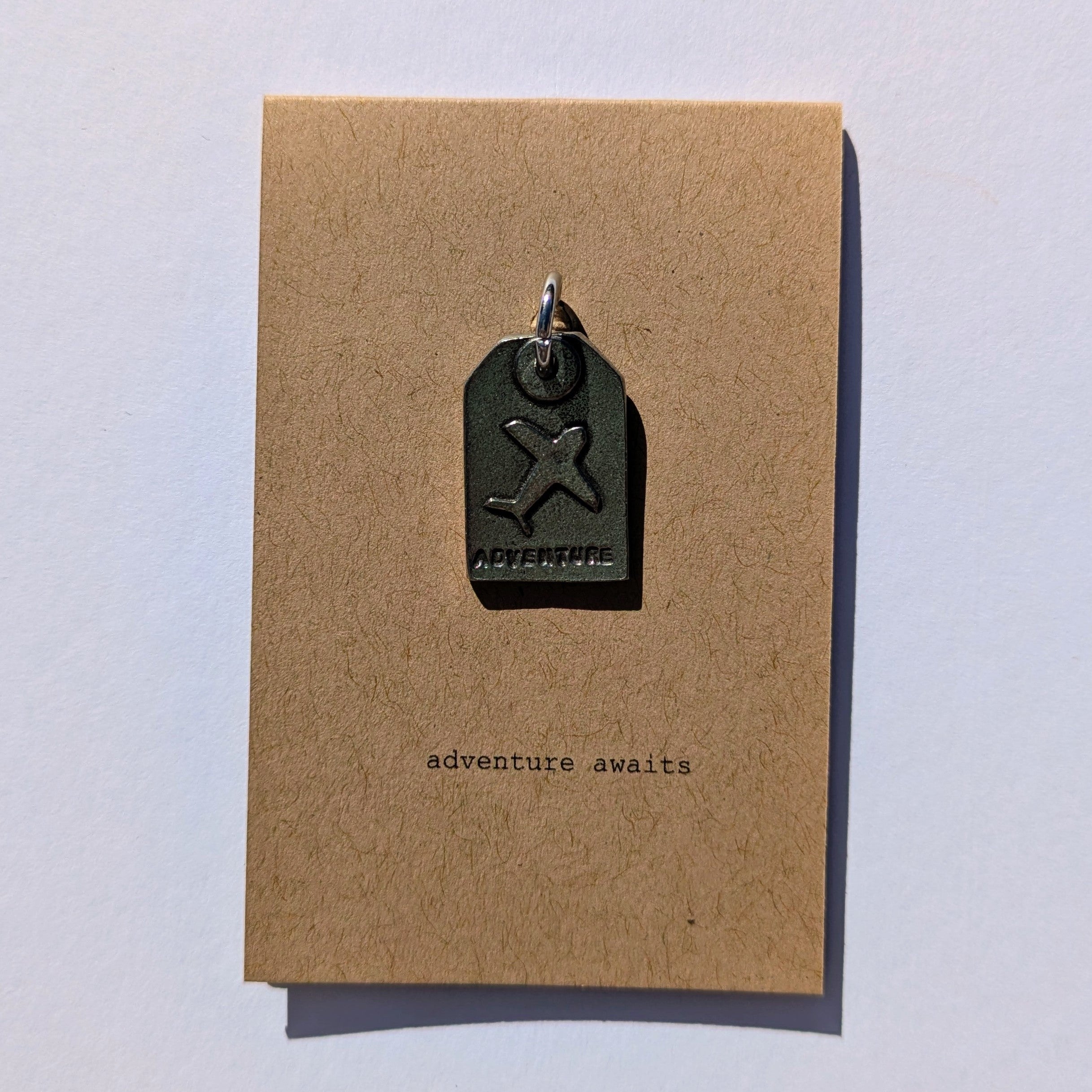 Metal plane tag shaped pendant on a brown card with 'adventure awaits' text