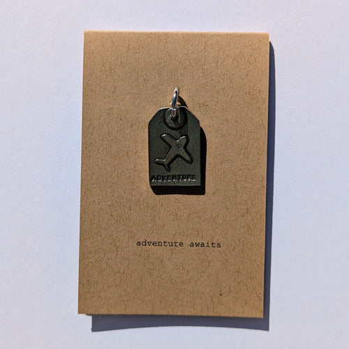 Metal plane tag shaped pendant on a brown card with 'adventure awaits' text